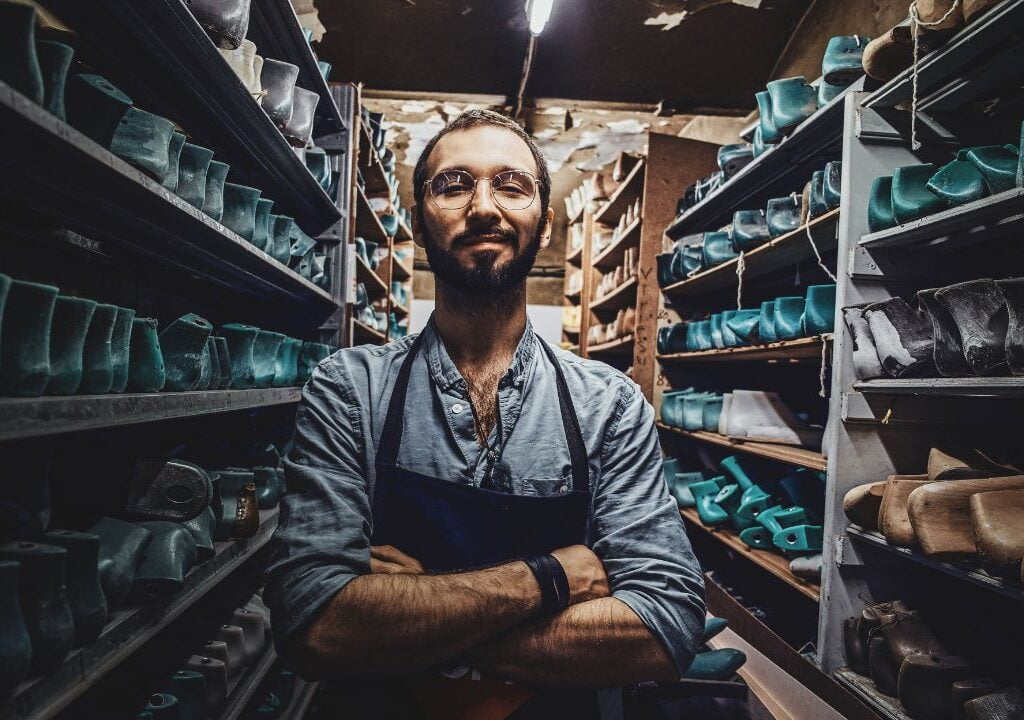 Bearded young cobbler wearing spectacles standing with hand folded in a storage surrounded with shoe form